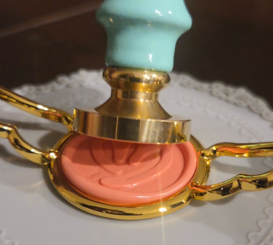 A coral wax seal in a gold stamp beside a mint-green handle.